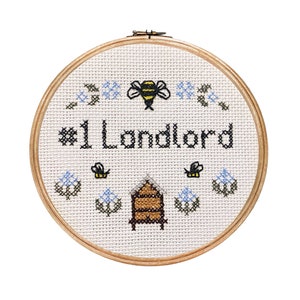 May include: A white embroidery hoop with a beige fabric background. The fabric has a cross-stitch design of a beehive, bees, flowers, and the text "#1 Landlord".