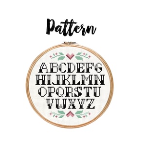 May include: A wooden embroidery hoop with a white fabric background and black letters spelling out the alphabet. The letters are surrounded by small green leaves and pink hearts.