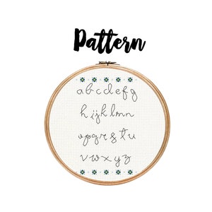 Puede incluir: Un aro de bordado blanco con un marco de madera y un patrón de punto de cruz del alfabeto en cursiva. Las letras están cosidas con hilo negro y el aro está decorado con pequeñas flores azules y amarillas.