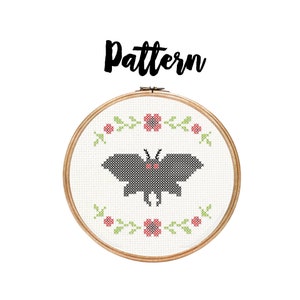 May include: A cross-stitch pattern featuring a black moth with red eyes, surrounded by a wreath of green leaves and pink flowers. The pattern is framed in a wooden hoop.