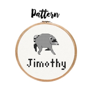 May include: A circular wooden embroidery hoop containing white Aida fabric with a cross-stitch design of a grey, pixelated, four-legged creature. Below the creature, the name Jimothy is stitched in black, blocky, pixel-style lettering.