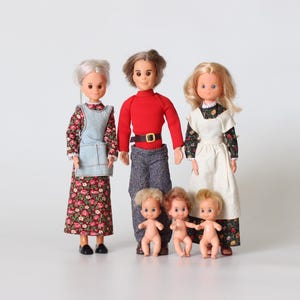 May include: A family of vintage dolls, including a grandmother in a floral dress and denim apron, a father in a red sweater, and a mother in a white dress. Three small baby dolls complete the family. The dolls have painted features and rooted hair.