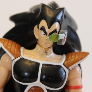 May include: A close-up of a Dragon Ball Z action figure. The figure has black spiky hair, a tan complexion, and a black and white chest plate. It wears a green-lensed scouter and orange shoulder pads. A red band is on the arm.
