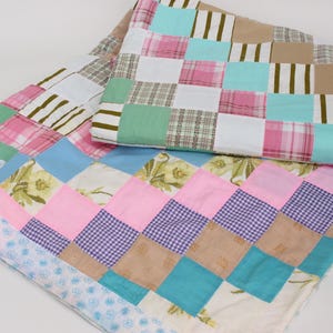 May include: Three patchwork quilts in various colours and patterns. The quilts feature squares of pink, blue, green, and tan fabrics, some with floral, plaid, and striped designs. The quilts are folded and stacked on a white surface.