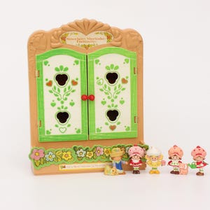 May include: A vintage Strawberry Shortcake toy set. It includes a tan wardrobe with green and white doors, and several small character figurines. The wardrobe has a floral base and the text "Strawberry Shortcake Fraisinette".