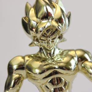 May include: A gold-colored figurine of a muscular anime character with spiky hair. The figure has a detailed face and is posed with arms slightly outstretched. The overall design suggests a superhero or warrior theme.