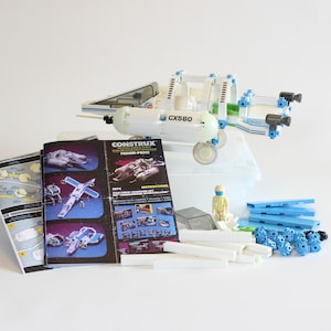 May include: A white and blue toy spaceship with the text "CX580" on the side. The spaceship is made of plastic and has a clear plastic dome on top. The spaceship is surrounded by instructions and extra pieces for the toy.