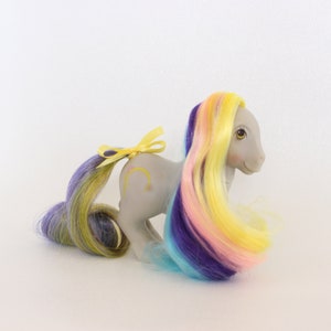 May include: A gray My Little Pony with a yellow ribbon in its tail. The pony has long, multi-colored hair with streaks of yellow, pink, blue, and purple.