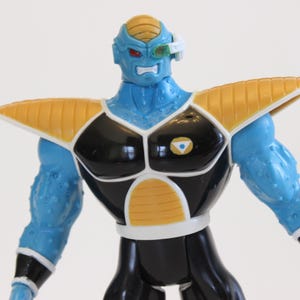 May include: A blue action figure with yellow and black armor. The figure has a blue head with a yellow crest and red eyes. It wears a white visor and has a muscular build. The figure is a collectible item.