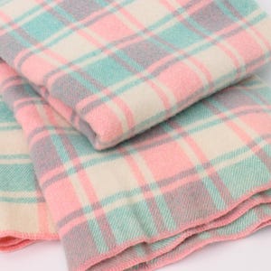 Peut inclure: Une couverture pliée avec un motif à carreaux aux couleurs pastel. La couverture présente un motif répétitif de carrés roses, bleu clair, gris et crème. Les bords sont finis avec une bordure rose. La couverture semble douce et confortable.