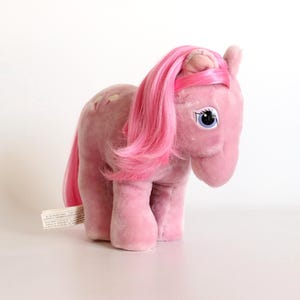 May include: A pink plush toy pony with a pink mane and tail. The toy has blue eyes and a tag attached to its side. The pony is standing on a white surface.