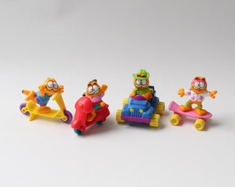 May include: Four Garfield character figurines on various vehicles. The figurines are orange with black stripes and white eyes. Vehicles include a yellow scooter, red scooter, blue jeep, and pink skateboard.