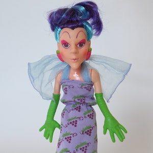 Peut inclure: Une poupée aux cheveux violets, des gants verts et une robe violette à motif de raisin. La poupée a une coiffure bleu et violet et un maquillage rose et vert.