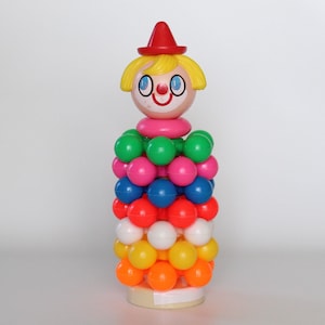 May include: A vintage toy clown with a red hat, yellow hair, and a pink neck ring. The clown's body is made of colourful plastic balls in various colours, including green, pink, blue, orange, white, and yellow. The toy is standing on a white base.