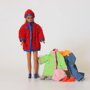 May include: A doll wearing a red coat and matching hat with blue trim. Additional clothing items include a green coat, orange shirt, blue jeans, pink hat, and orange shoes. The doll is standing on a white surface.