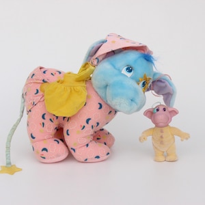 May include: A plush toy elephant with a blue head and pink body, wearing a pink hat and yellow saddle. A small, yellow figurine stands nearby. The elephant's body is patterned with blue and white shapes.