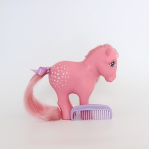 May include: A pink toy pony with white spots on its side, a pink tail, and a purple bow. A matching purple comb rests in front of the pony. The pony has blue eyes and a pink mane.