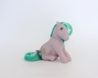 May include: A purple My Little Pony with green hair and a green and purple bow. The pony has green clover-shaped markings on its side.