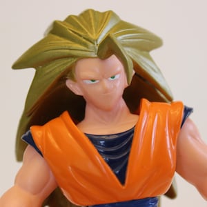 May include: A close-up of a plastic action figure of a character with spiky, golden hair. The figure wears an orange and blue outfit. The character has a determined expression with green eyes. The figure is a collectible item.