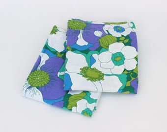 May include: Two folded fabric pieces with a floral pattern. The design features large white flowers with green centers, purple petals, and teal and green leaves on a white background.