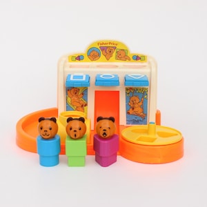 Peut inclure: Un jouet Fisher-Price vintage sur le thème des ours. Le jouet a une structure crème avec des accents bleus, orange et jaunes. Trois figurines en forme d'ours se tiennent devant le jouet, chacune avec une base de couleur différente.
