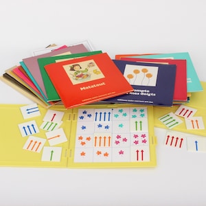Peut inclure: Un jeu éducatif jaune avec une pile de livres colorés et de petites cartes. Les livres ont des titres comme "Matatout" et "Compte mes doigts". Les cartes présentent des flèches et des motifs d'étoiles.