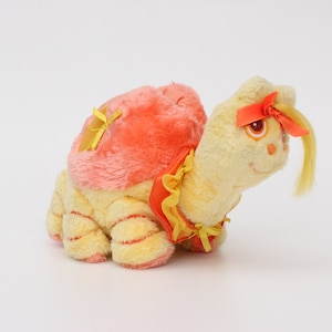 May include: A yellow plush turtle with a pink and orange shell and a yellow bow. The turtle has a yellow bow on its head and a yellow bow on its shell.