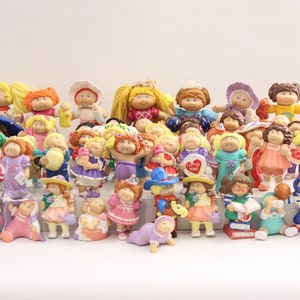 Peut inclure: Une collection de poupées Cabbage Patch Kids dans différentes poses et tenues. Les poupées sont en plastique et ont des visages peints. Certaines poupées tiennent des accessoires, comme un arrosoir, un livre et un ballon de football. Les poupées sont toutes de couleurs différentes et ont des coiffures différentes.