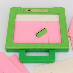 Peut inclure: Un jouet en plastique vert avec une poignée et une fenêtre transparente. Le jouet a une surface rose et blanche à l'intérieur de la fenêtre. Il y a aussi plusieurs tuiles en plastique rose et blanche.