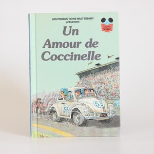 May include: A vintage book titled "Un Amour de Coccinelle" by Les Productions Walt Disney. The cover features a light green background with a drawing of a white Volkswagen Beetle racing on a track, with the number 53 on the side.