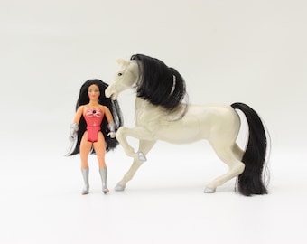 May include: A vintage doll with long black hair, wearing a red and silver outfit, stands next to a white toy horse with a black mane and tail. The doll has silver boots and gauntlets. The horse is in a trotting pose.