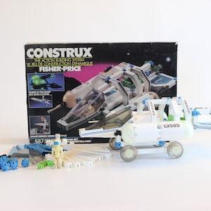 May include: A vintage Constux building set by Fisher-Price, featuring a space theme. The set includes a spaceship, a CX580 vehicle, and various building components in white, blue, and green. The box art displays the set's features.