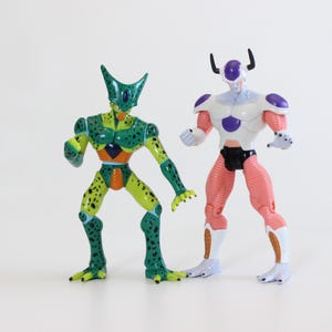May include: Two action figures of characters from the Dragon Ball Z anime series. One figure is green and yellow with black spots, the other is white, purple, and pink. Both figures are standing with their arms at their sides.