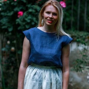 May include: A woman wearing a blue linen top with a white linen skirt. She is standing in front of a bush of pink roses.