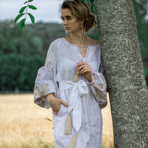 Puede incluir: Una mujer con un vestido blanco de lino con detalles bordados y un cintur&oacute;n blanco. El vestido tiene borlas en el cintur&oacute;n y las mangas. Est&aacute; de pie junto a un &aacute;rbol.