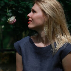 May include: A woman with long blonde hair wearing a dark blue linen top and long, white, shell-shaped earrings. She is standing in front of a lush green bush with pink roses.