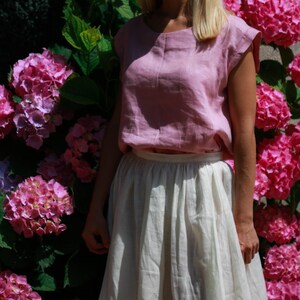 May include: A woman wearing a light pink linen top with a white linen skirt. She is standing in front of a wall of pink hydrangeas.