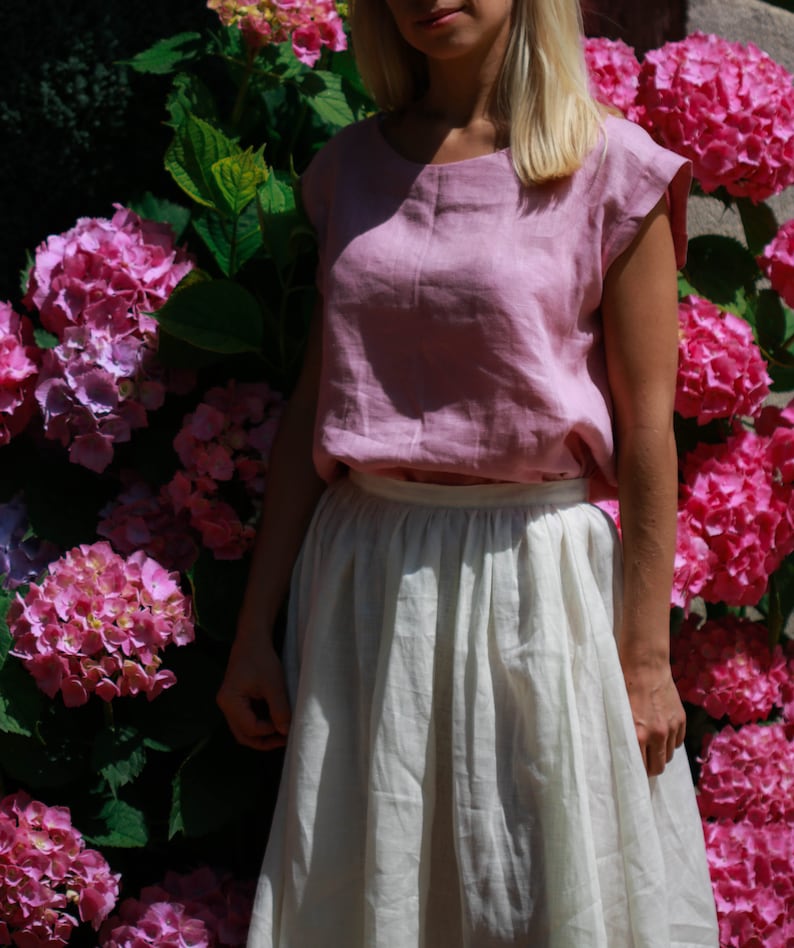 May include: A woman wearing a light pink linen top with a white linen skirt. She is standing in front of a wall of pink hydrangeas.
