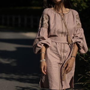 K&ouml;nnte beinhalten: Eine Frau tr&auml;gt ein langes, hellrosa Leinenkleid mit Stickereien und einem braunen G&uuml;rtel. Das Kleid hat lange &Auml;rmel mit einer gerafften Puff&auml;rmeln an der Schulter und einem Quasten-Detail an der Taille.