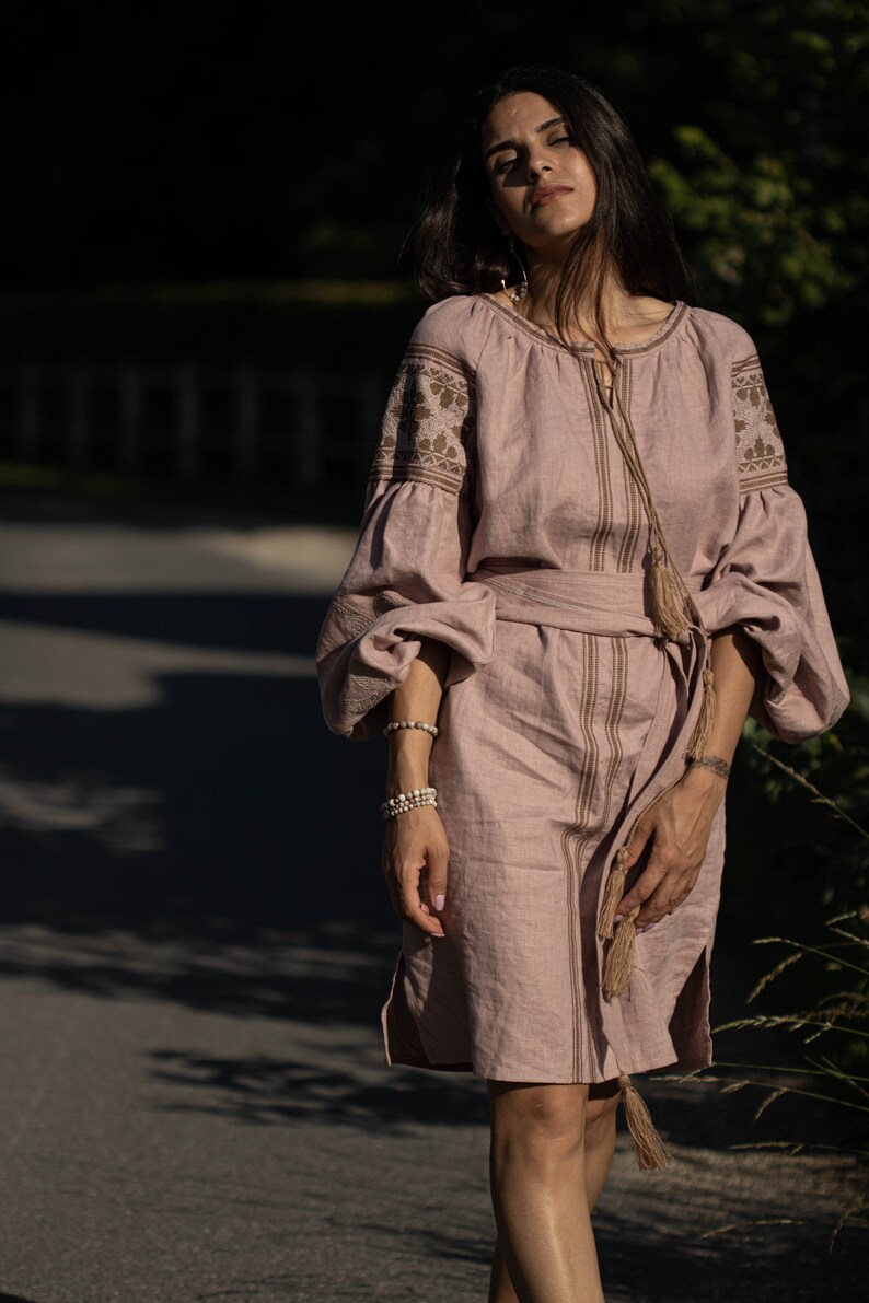 K&ouml;nnte beinhalten: Eine Frau tr&auml;gt ein langes, hellrosa Leinenkleid mit Stickereien und einem braunen G&uuml;rtel. Das Kleid hat lange &Auml;rmel mit einer gerafften Puff&auml;rmeln an der Schulter und einem Quasten-Detail an der Taille.