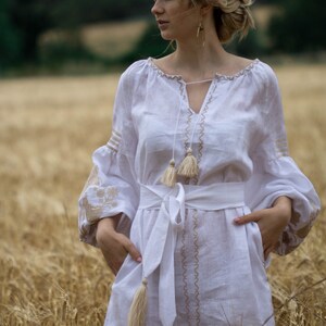 Puede incluir: Una mujer con un vestido blanco de lino con detalles bordados y borlas se encuentra en un campo de trigo. El vestido tiene un escote en V y una cinta blanca atada a la cintura.