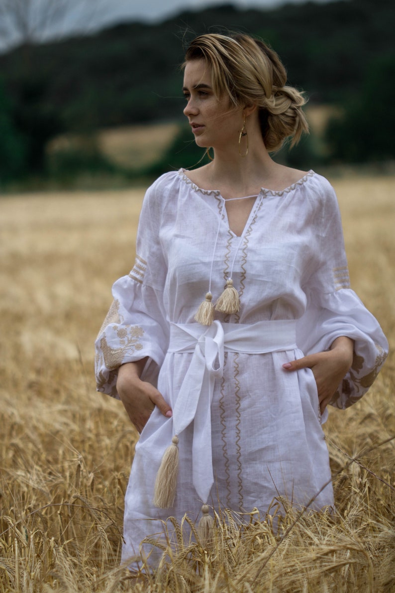 Puede incluir: Una mujer con un vestido blanco de lino con detalles bordados y borlas se encuentra en un campo de trigo. El vestido tiene un escote en V y una cinta blanca atada a la cintura.
