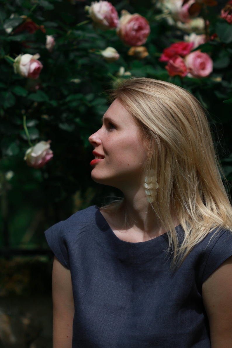 May include: A woman with long blonde hair wearing a dark blue linen top and long, white, shell-shaped earrings. She is standing in front of a lush green bush with pink roses.
