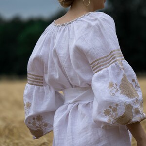 Puede incluir: Una blusa de lino blanca con dise&ntilde;os florales bordados en color marr&oacute;n claro. La blusa tiene un escote redondo fruncido y mangas largas con un pu&ntilde;o fruncido. La blusa est&aacute; ce&ntilde;ida a la cintura con una faja blanca.