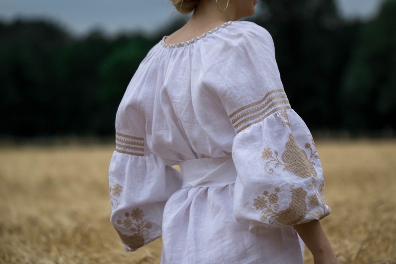 Puede incluir: Una blusa de lino blanca con dise&ntilde;os florales bordados en color marr&oacute;n claro. La blusa tiene un escote redondo fruncido y mangas largas con un pu&ntilde;o fruncido. La blusa est&aacute; ce&ntilde;ida a la cintura con una faja blanca.