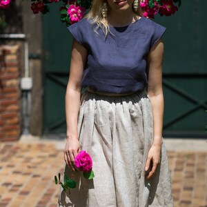 May include: A woman wearing a navy blue linen top and a beige linen skirt. She is holding a pink rose in her hand. She is standing in front of a brick wall and a green door.