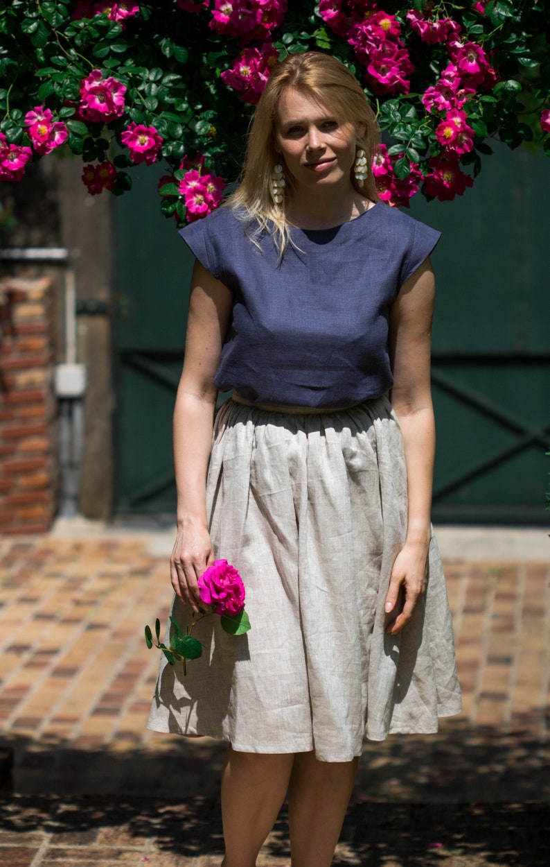 May include: A woman wearing a navy blue linen top and a beige linen skirt. She is holding a pink rose in her hand. She is standing in front of a brick wall and a green door.