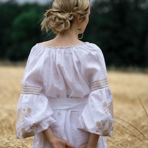 Puede incluir: Una mujer con un vestido blanco de lino con detalles bordados se encuentra en un campo de hierba alta. El vestido tiene un escote drapeado y mangas largas con pu&ntilde;os bordados. El cabello de la mujer est&aacute; recogido en un mo&ntilde;o trenzado.