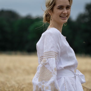 Puede incluir: Una mujer con un vestido blanco de lino con detalles bordados se encuentra en un campo de trigo. El vestido tiene una faja blanca atada a la cintura y el bordado es de color beige. La mujer sonr&iacute;e y mira por encima del hombro.