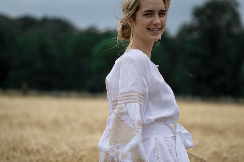 Puede incluir: Una mujer con un vestido blanco de lino con detalles bordados se encuentra en un campo de trigo. El vestido tiene una faja blanca atada a la cintura y el bordado es de color beige. La mujer sonr&iacute;e y mira por encima del hombro.